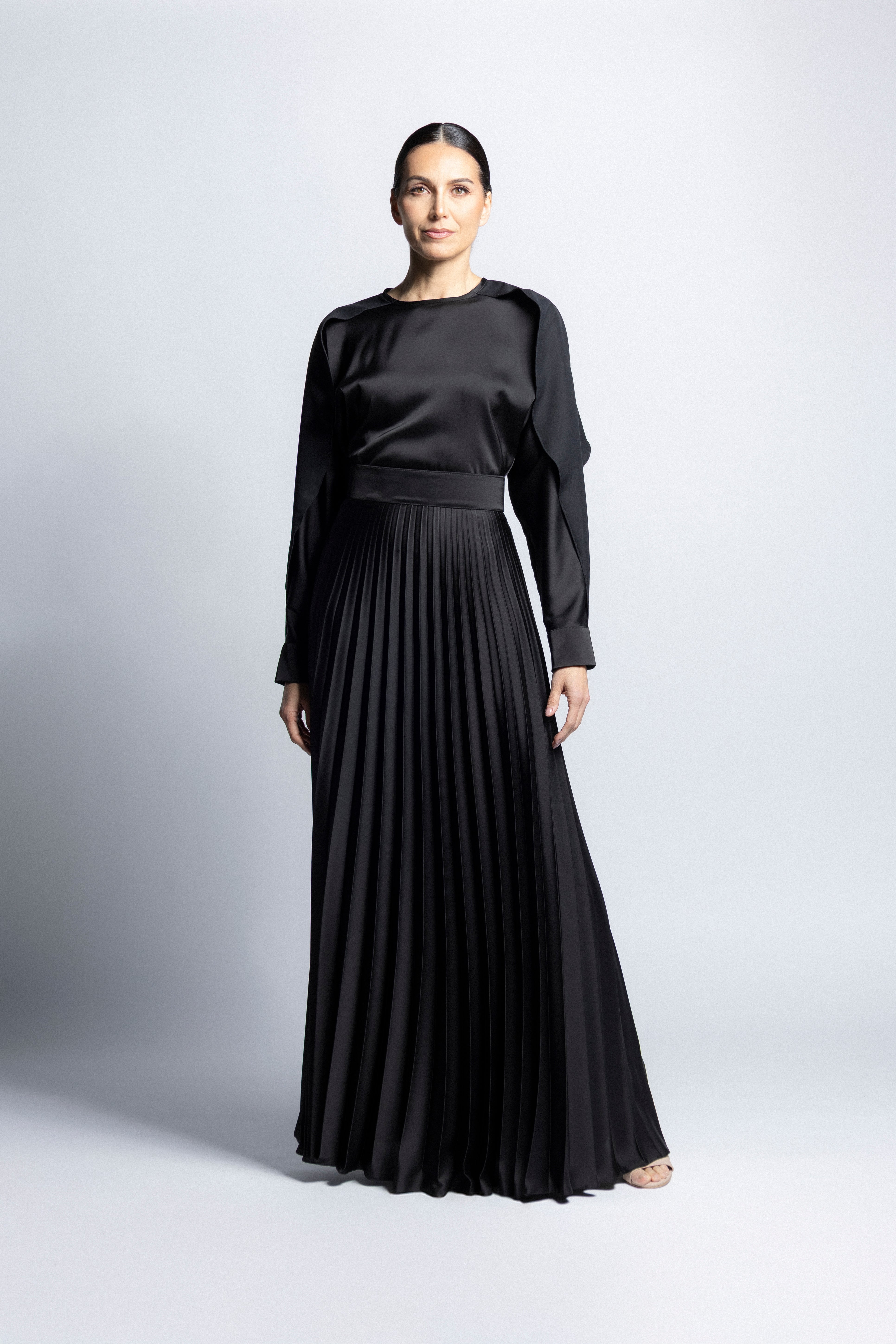 Vestido largo de satín en color negro  con torso de cuello redondo, mangas largas con puños detallados y falda larga plisada.