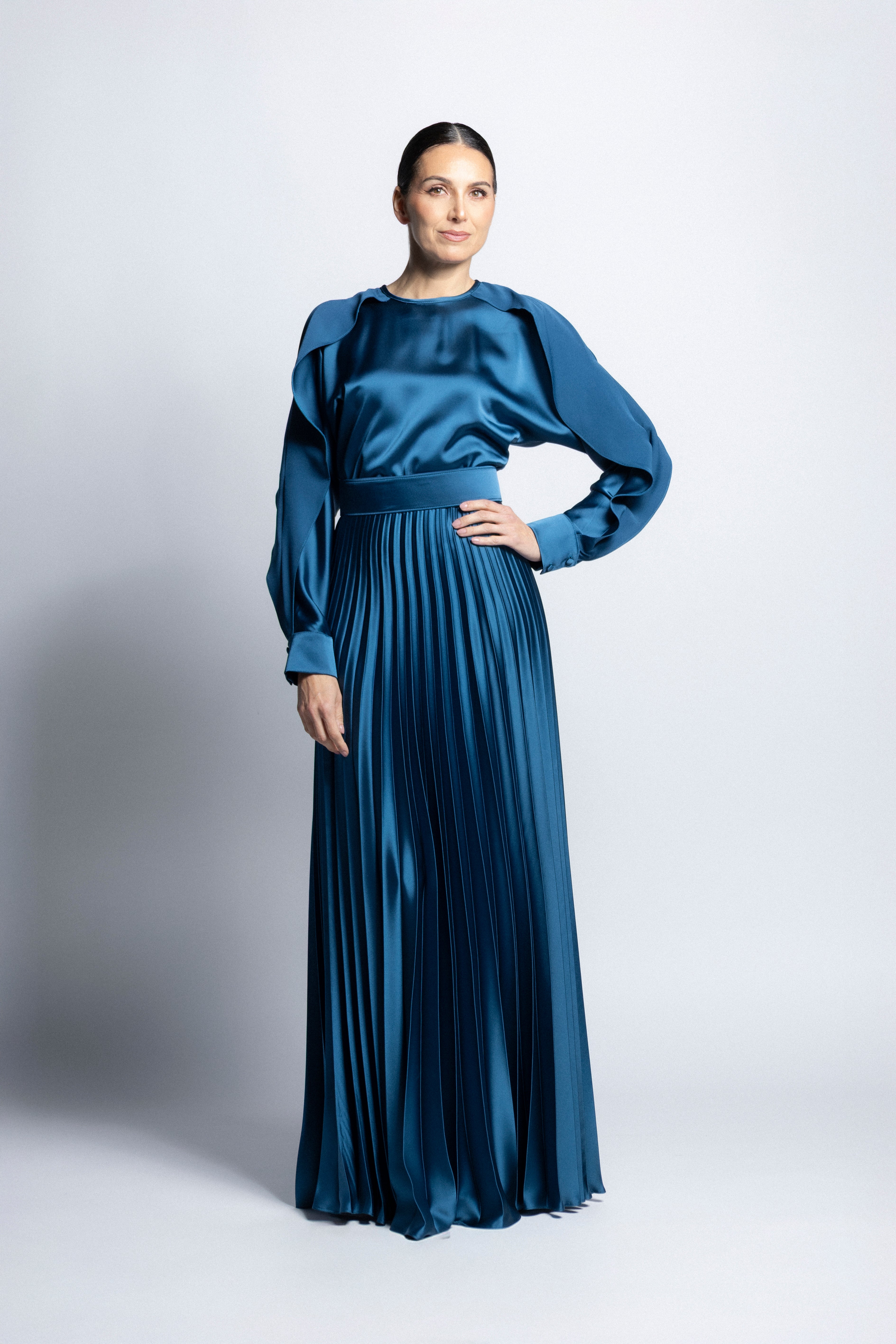 Vestido largo de satín en color azul petróleo con torso de cuello redondo, mangas largas con puños detallados y falda larga plisada.