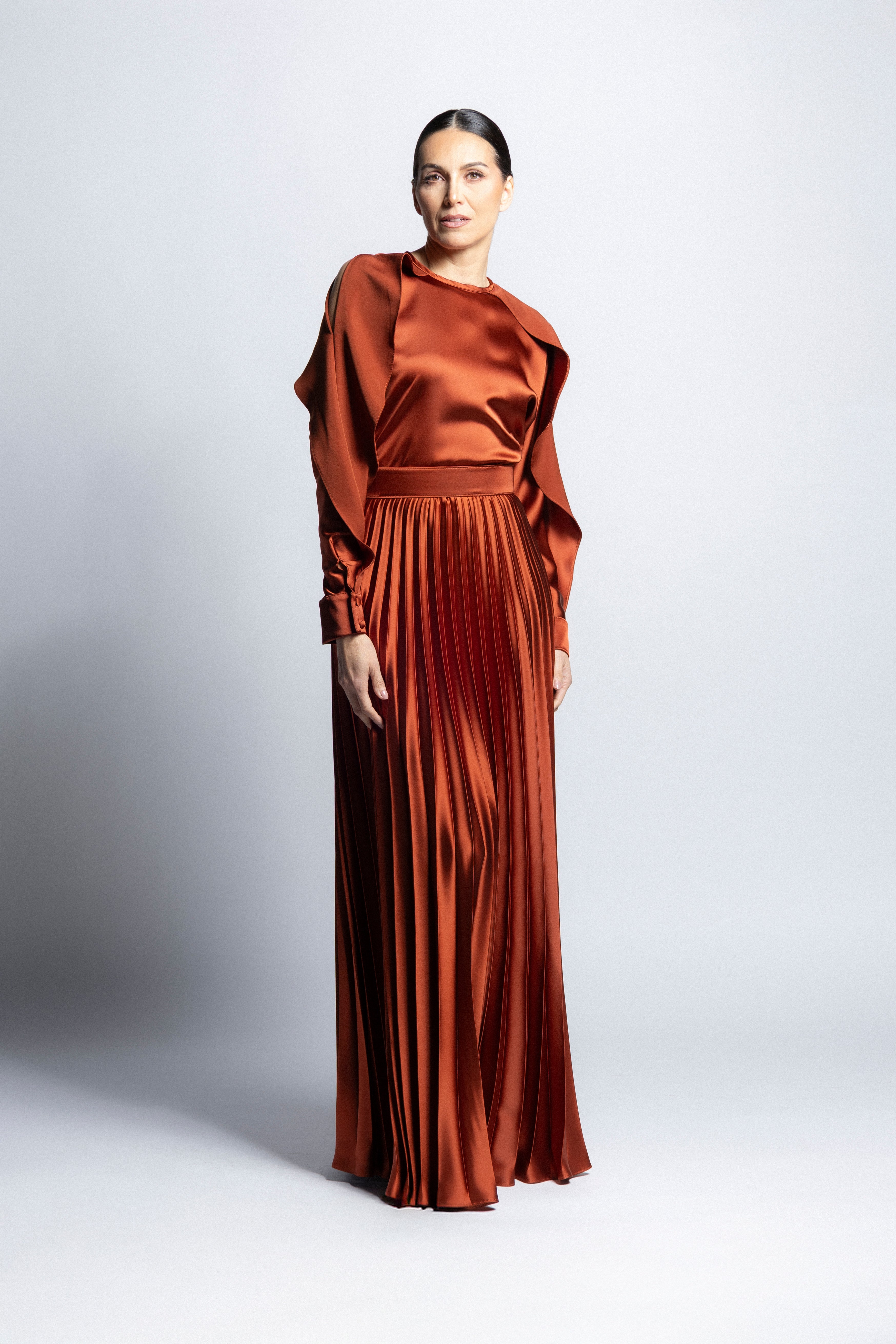 Vestido largo de satín en color cobrecon torso de cuello redondo, mangas largas con puños detallados y falda larga plisada.